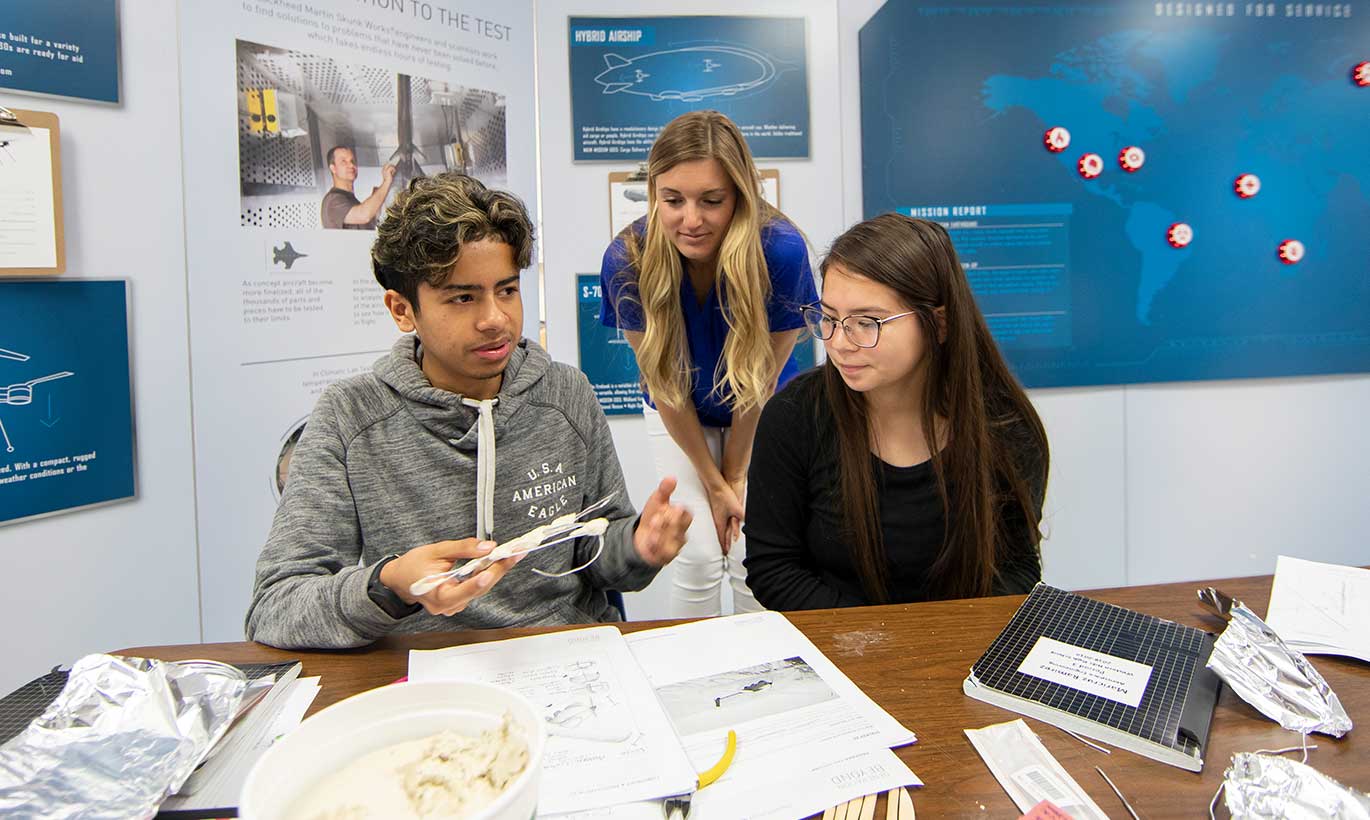 Three young people sit at a table with papers, books, and research in front of them