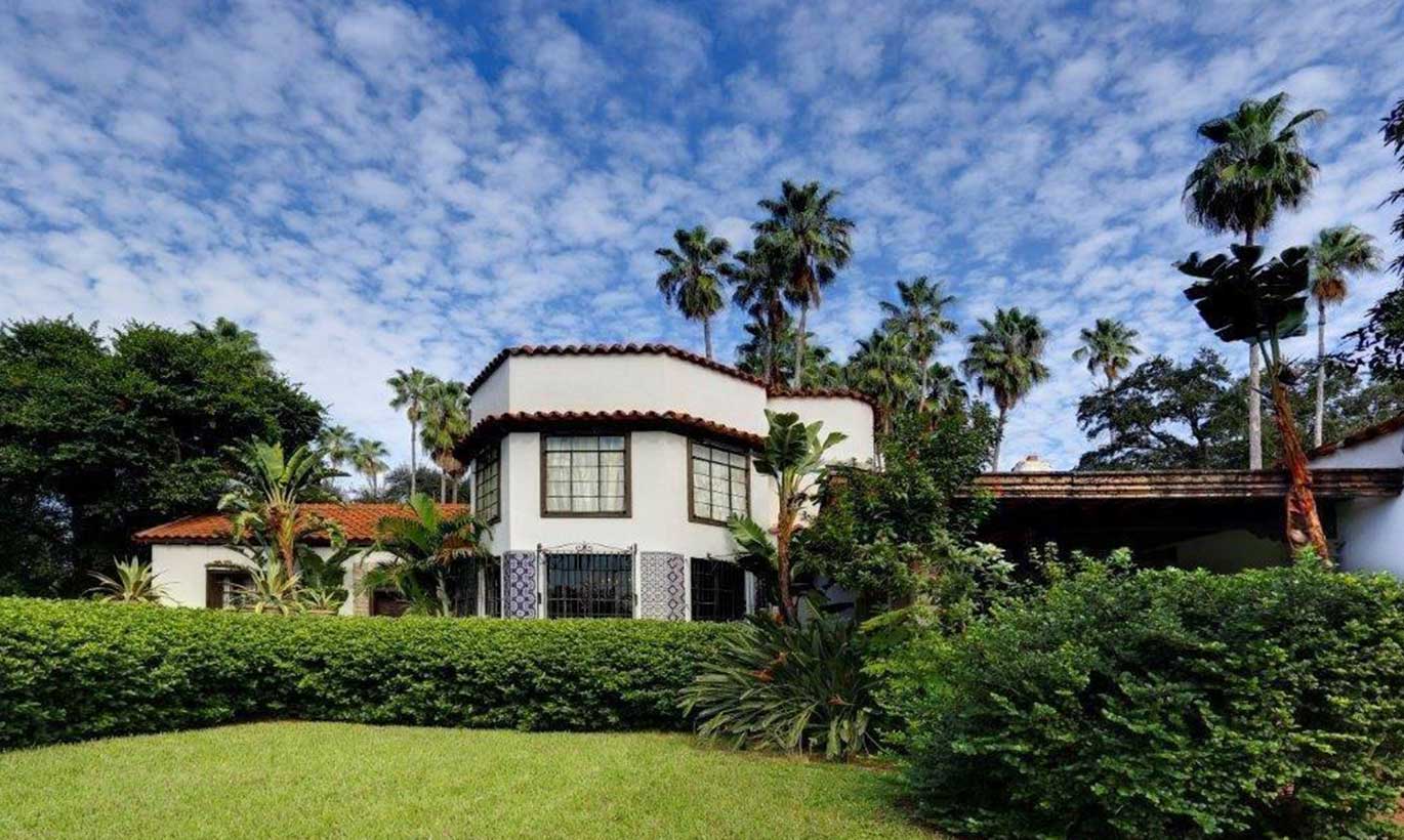A white Spanish style house with an orange terracotta roof is surrounded by palm trees