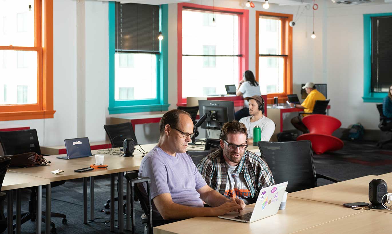 Two men work on a laptop computer with brightly colored window frames behind them and other workers in the background at the Geekdom Coworking Space in San Antonio, Texas.