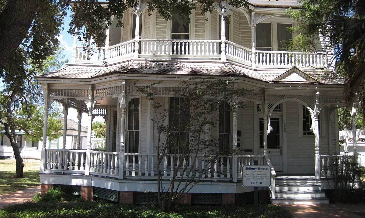 The Sidbury House, a historical building in Corpus Christi, has light yellow siding and a white porch, surrounded by trees.