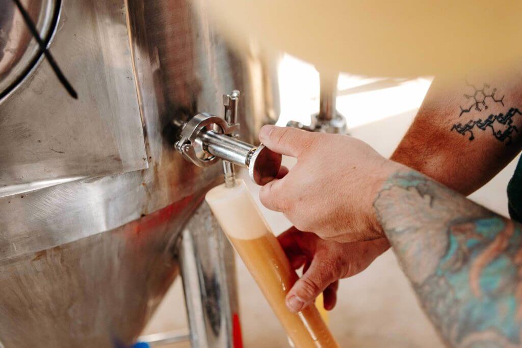 A man's hands with colorful tattoos pour out a beer from a tap into a tall, skinny glass.