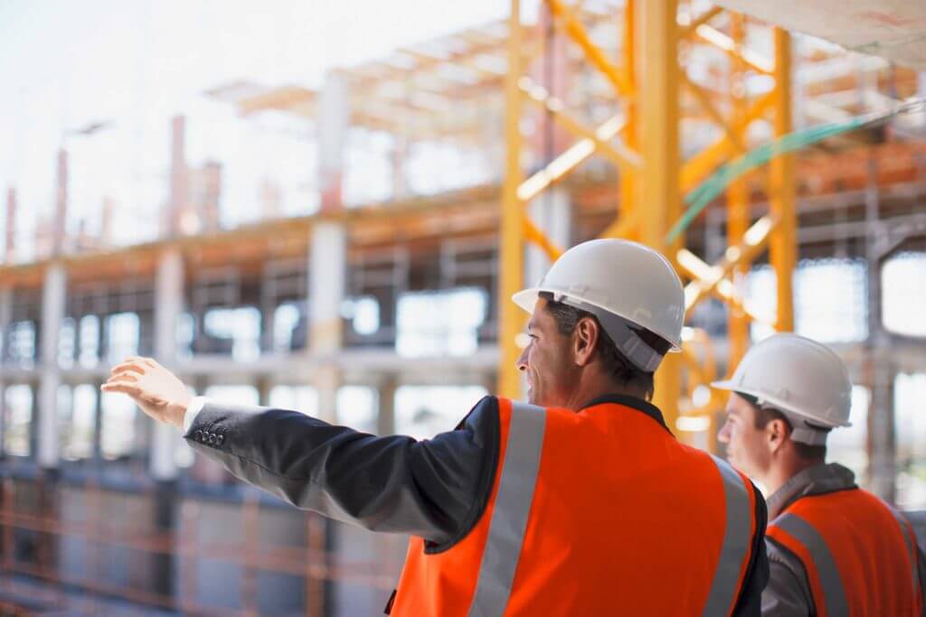 Two men in hard hats and orange safety vests gesture toward their work site.