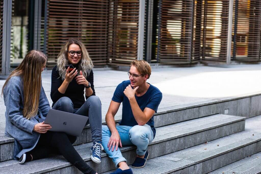 Three college students sit outside on the steps of a building with their laptops.