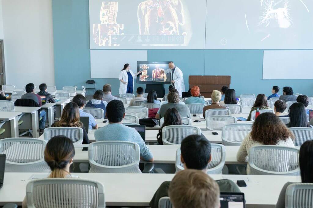 College students sit at long white tables in a lecture hall while two professors in white lab coats present a screen showing human anatomy in front of the classroom.