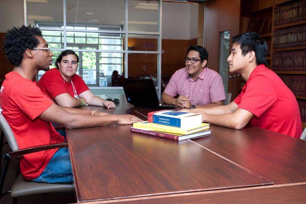 Four students wearing red shirts sit at a table having a discussion at the University of Houston.