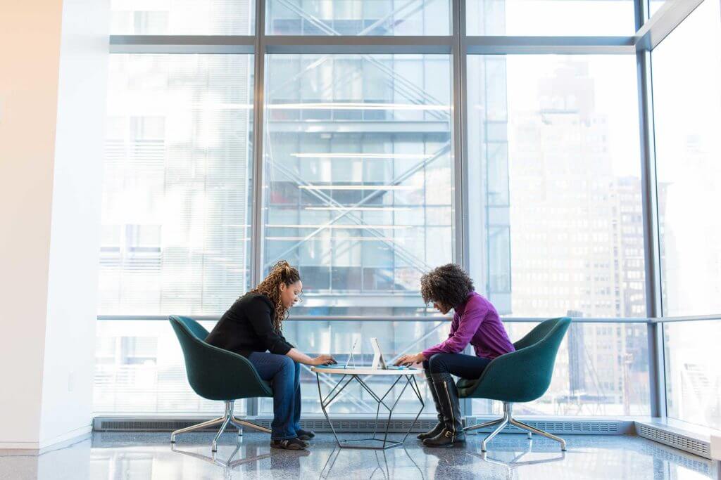 Two women sitting on green padded chairs in an office workspace typing on their laptop computers