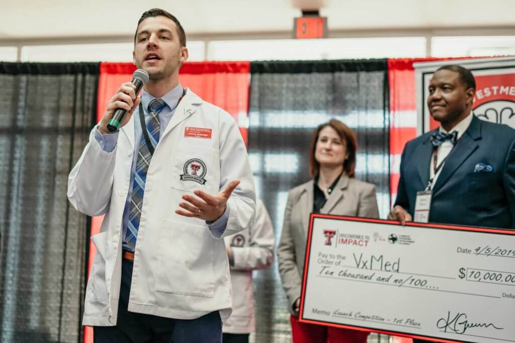 A man wearing a white lab coat speaks into a microphone while a man behind him holds a large cardboard check with a donation amount of $10,000.