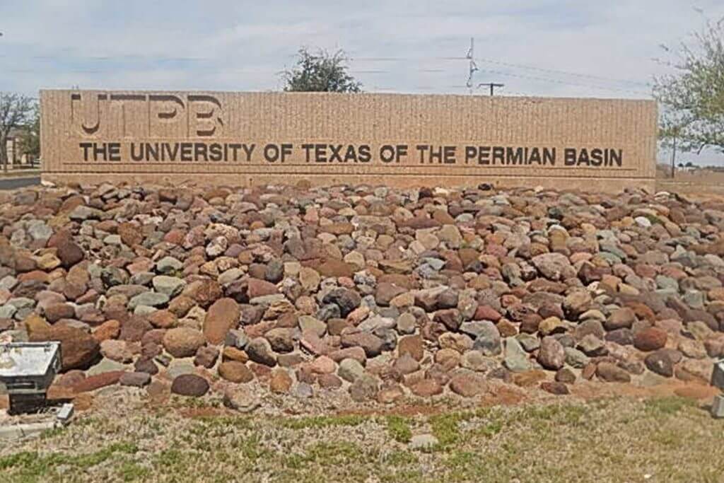 A large beige sign reading "The University of Texas of the Permian Basin" sits atop a large pile of decorative rocks.