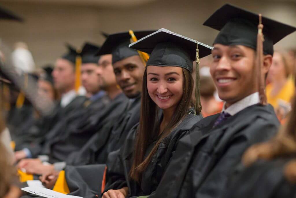 Three college graduates wearing black gowns and black caps with yellow tassels smile at the camera.