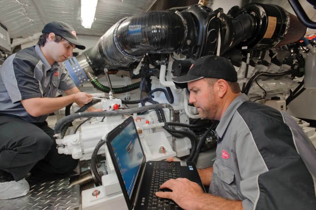 Two workers in gray uniforms use a laptop computer and metal tools to work on a product.